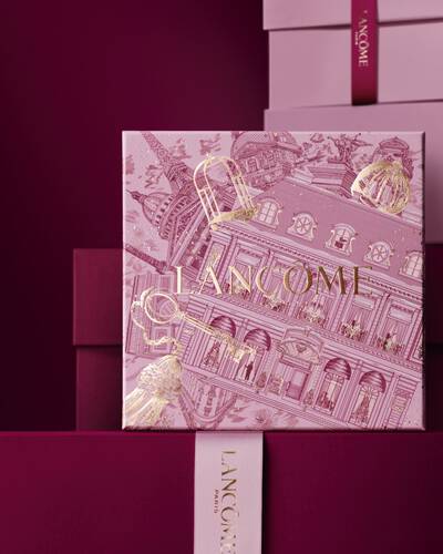 Image of pink Lancome holiday gift box standing in front of a pile of pink and dark red gift boxes, set against a dark red background.