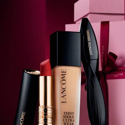 An elegant still life of Lancome's L'Absolu Rouge matte lipstick, Teint Idole Ultra Wear foundation, and Hypnose Drama mascara set against a luxurious dark red holiday background.