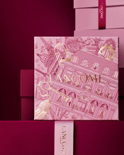 Image of pink Lancome holiday gift box standing in front of a pile of pink and dark red gift boxes, set against a dark red background.