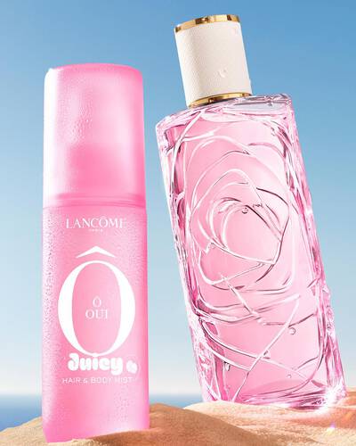 Three colorful spray bottles, one yellow, one pink, and one green, standing on a sandy beach next to a pink striped towel. The ocean is in the background under a clear blue sky.