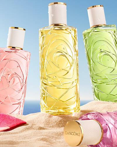 Two pink bottles, one perfume transparent glass bottle with a textured design and one spray bottle, standing on a sandy beach. A folded pink striped beach towel lies next to the bottles. In the background, there is a clear blue sky and the ocean.