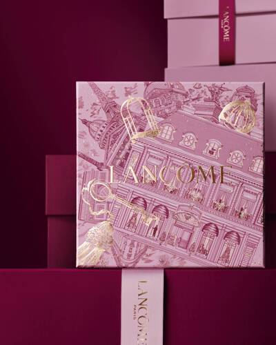 Image of pink Lancome holiday gift box standing in front of a pile of pink and dark red gift boxes, set against a dark red background.
