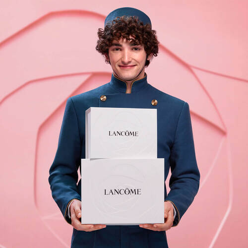 A smiling young man in a dark blue uniform holds two white boxes with the name "Lancome" displayed on them, against a soft pink background featuring a rose design.