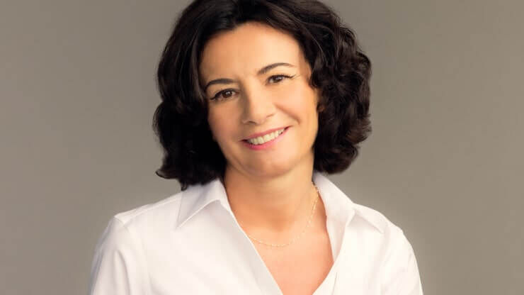 A woman with curly dark hair smiles warmly, wearing a white collared shirt against a neutral background.