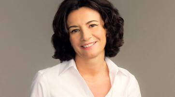 A woman with curly dark hair smiles warmly, wearing a white collared shirt against a neutral background.