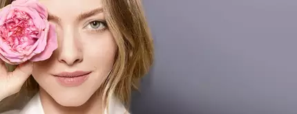 A woman with light brown hair holds a pink rose in front of her eye, smiling softly against a muted gray background.