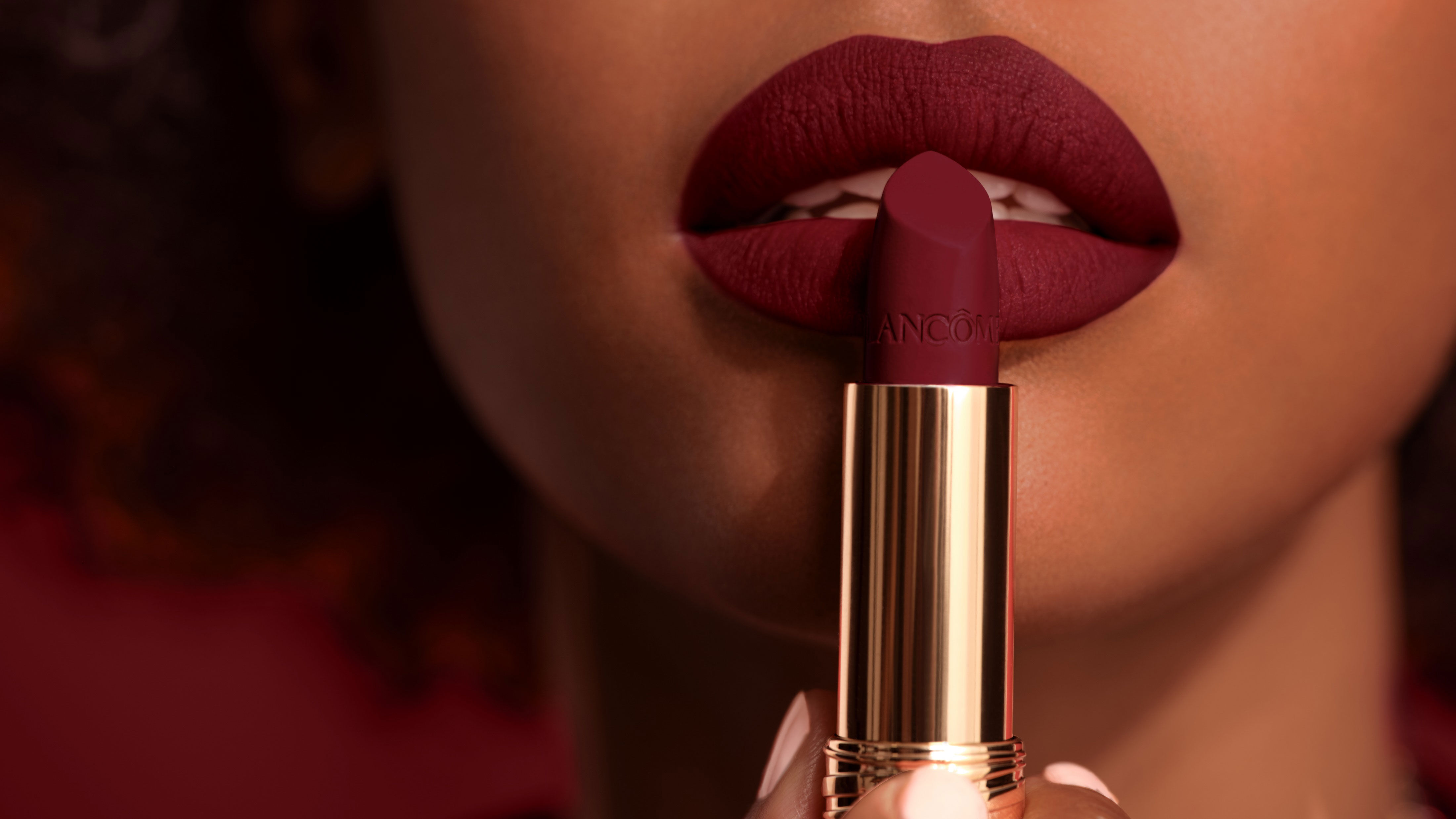Close-up of a person holding a tube of deep red lipstick near their lips, which are painted in the same color, showcasing a matte finish.