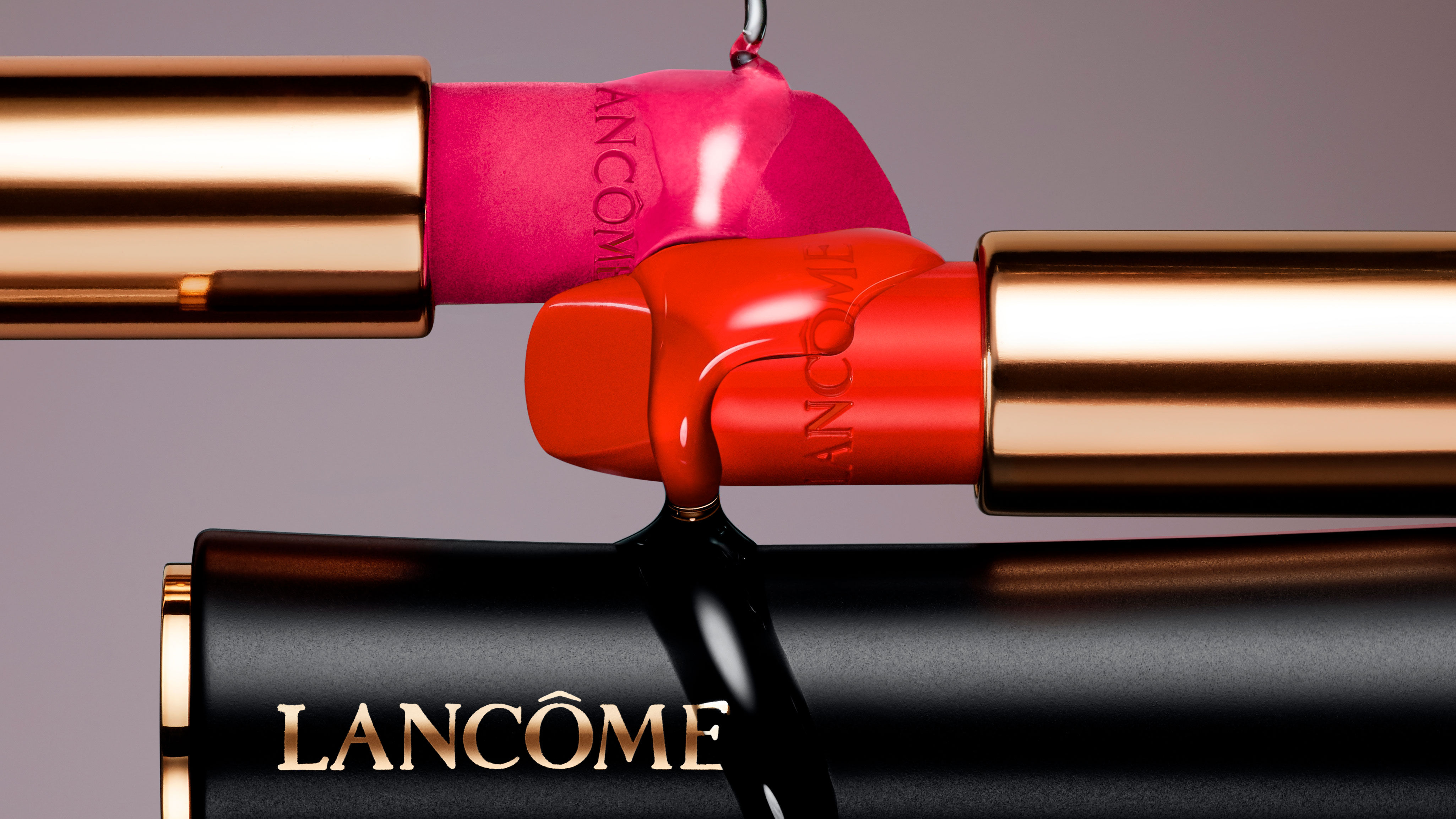 Three lipstick tubes in shades of pink and red are artistically arranged. The tubes have gold accents and the Lancome logo prominently displayed. The background is a soft gradient.