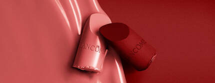 A close-up of two lipstick bullets, one in a light pink and the other in a deep red, against a smooth red background. The lipsticks have the brand name "Lancome" engraved on them.