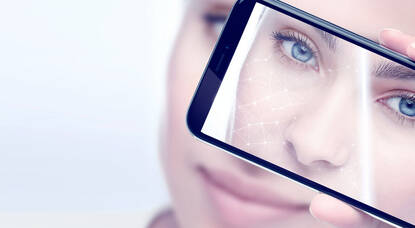 A close-up image of a person holding a smartphone up to their face, displaying a digitally enhanced reflection of their skin with bright lines and facial analysis features. The individual has blue eyes and a subtle smile.