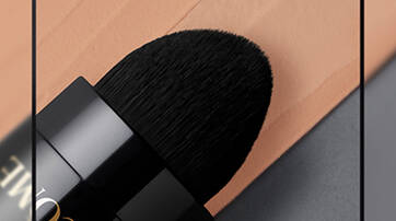 Close-up of a makeup brush with a soft, black bristle tip resting next to a swatch of peach-toned foundation on a gray background.