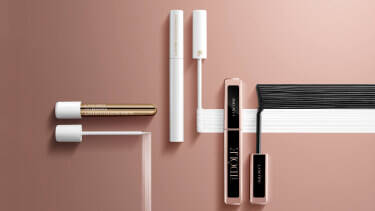 Flat lay of various Lancome mascara products arranged on a soft pink background, including a gold and white tube, a black tube, and a spoolie brush.