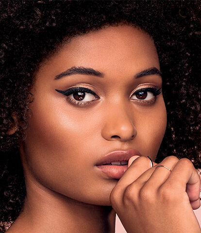 A close-up portrait of a woman with curly hair and striking makeup, showcasing a thoughtful expression against a soft, pastel background.