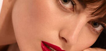 Close-up portrait of a woman with dark hair and full red lips, wearing a white blouse and a gold bracelet, gazing directly at the camera.