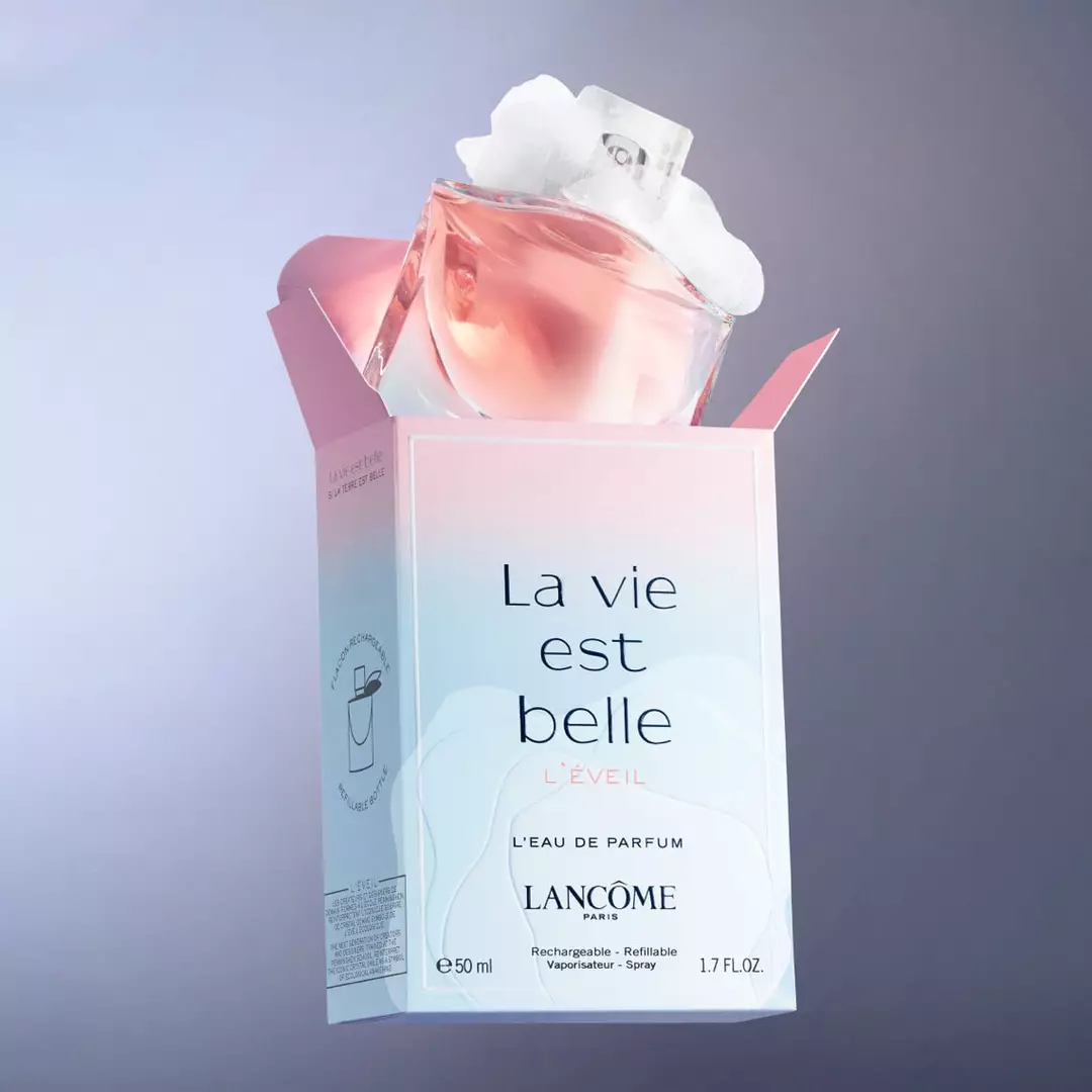 A bottle of Lancome's "La vie est belle L'eveil" perfume, featuring a soft pink liquid and a decorative cap, emerging from its pastel-colored box against a gradient background.