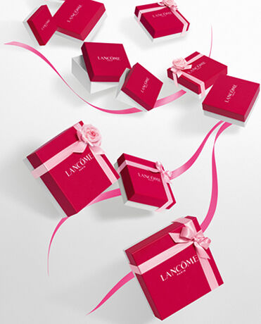 A collection of elegant red gift boxes with white accents, featuring the Lancome logo, arranged artistically on a light background, accompanied by pink ribbons and a flower.