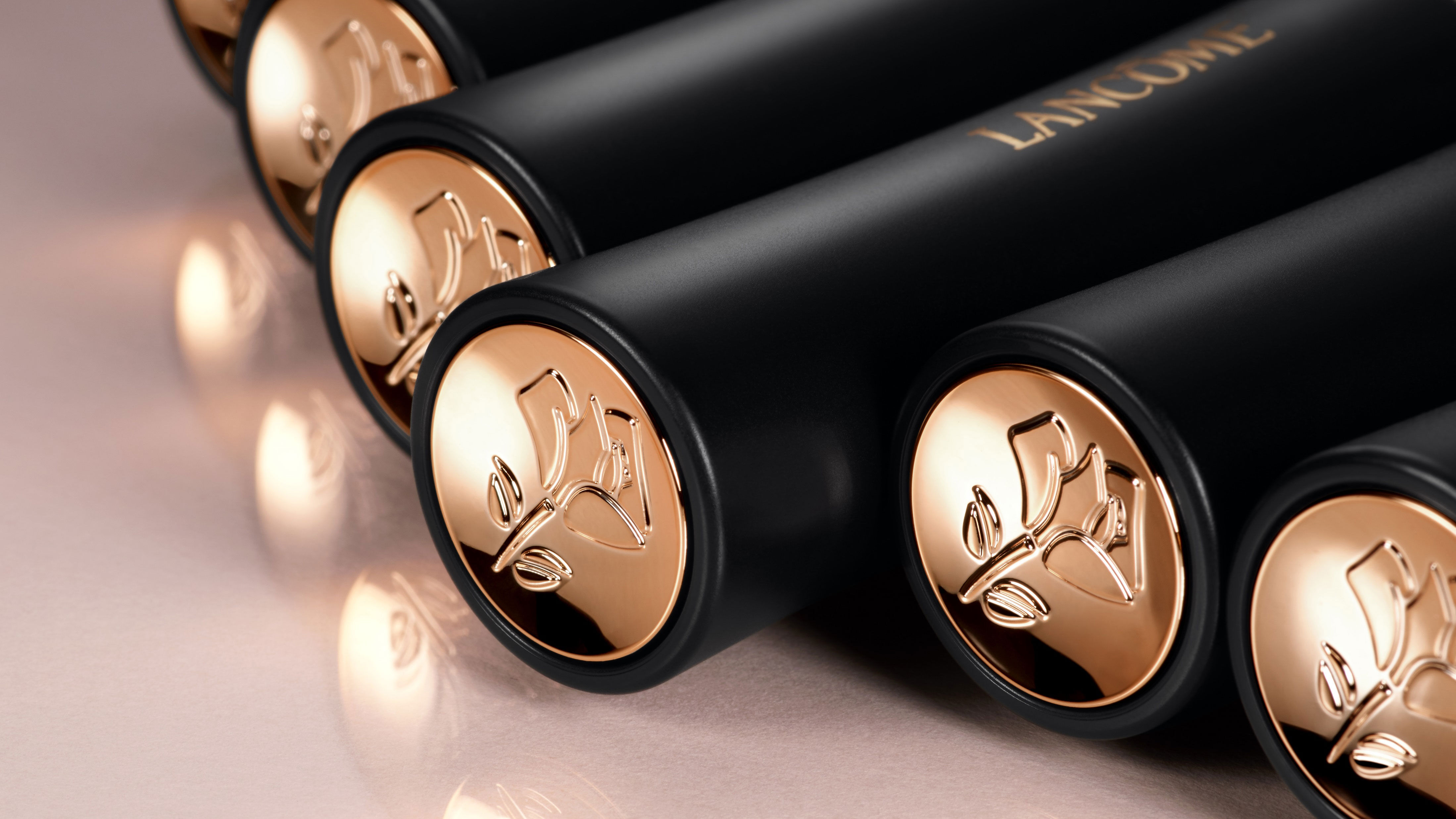 Close-up of black cylindrical tubes with gold caps, featuring an embossed floral design, arranged in a row. The branding "Lancome" is visible on the tubes. The surface reflects light, adding a glossy effect.