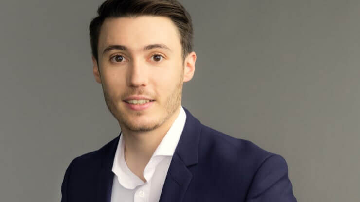 A young man with short dark hair, wearing a navy blazer over a white shirt, poses against a plain gray background. He is smiling and looking directly at the camera.