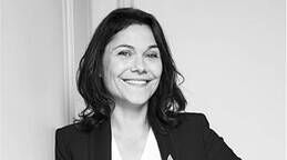 A smiling woman with dark, wavy hair, wearing a black blazer over a white top, stands against a light-colored wall.