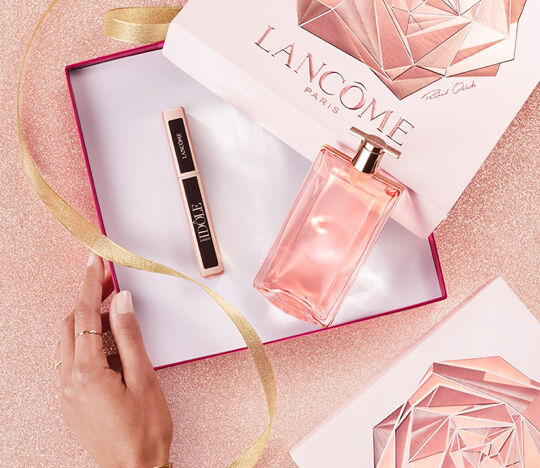 A hand rests on a pink surface near an open gift box containing a bottle of Lancome perfume and a mascara. The box features a geometric design and a pink ribbon is elegantly draped nearby.