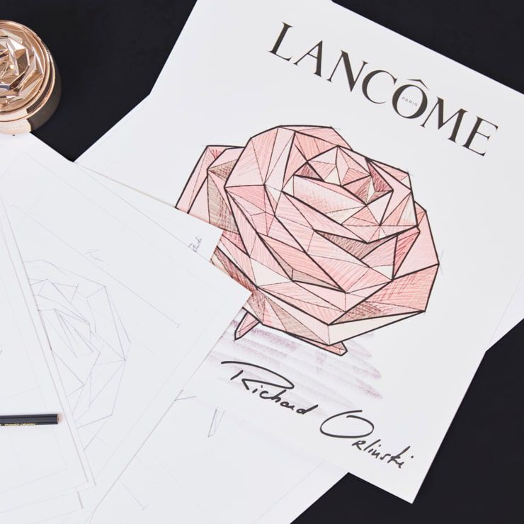 A close-up of a design featuring a geometric rose in shades of pink, accompanied by the text "LANCOME" and "Richard Orlinski." Surrounding sheets of paper and a pen are visible.