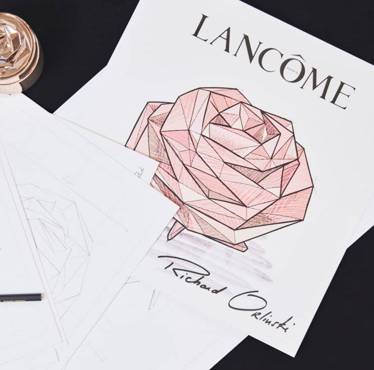 A close-up of a design featuring a geometric rose in shades of pink, accompanied by the text "LANCOME" and "Richard Orlinski." Surrounding sheets of paper and a pen are visible.