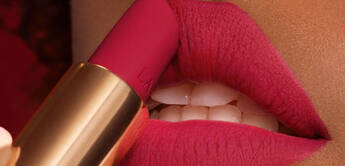 Close-up of a person applying dark pink lipstick to their lips, showcasing a smooth, matte finish. The background features a subtle red hue.