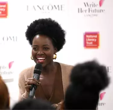 Lupita Nyong'o smiles while speaking into a microphone, with "LANCOME" and "Write Her Future" logos in the background.
