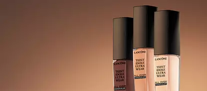 Three bottles of foundation are displayed against a warm, gradient background. Each bottle has a sleek black cap and features labels indicating the product name, "Extreme Wear."
