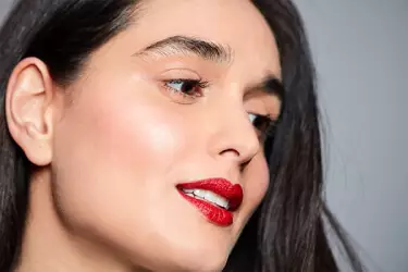 A close-up portrait of a woman with long, dark hair, smiling softly. She has a light makeup look with shimmery skin and bold red lipstick, set against a neutral gray background.