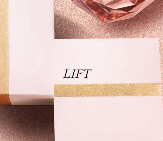 A close-up of two elegantly wrapped gift boxes in soft pink with gold accents, featuring the word "LIFT" on one of the boxes, set against a textured, warm background with a sparkling rose-colored crystal.