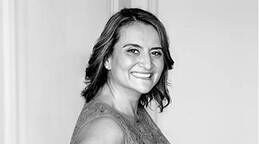 A smiling woman with shoulder-length, wavy hair is looking sideways, wearing a sleeveless top in a light-colored setting. The image is in black and white.