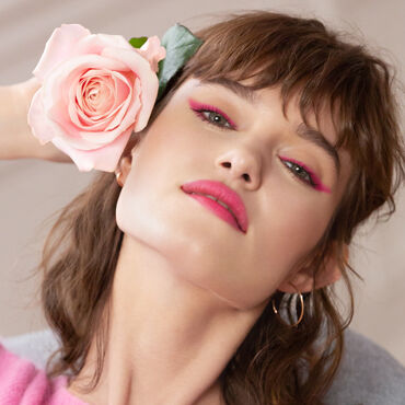 A woman with a softly styled hairstyle poses with a pink rose by her ear, featuring vibrant pink eye makeup and a rosy lip color, exuding a confident and elegant vibe.