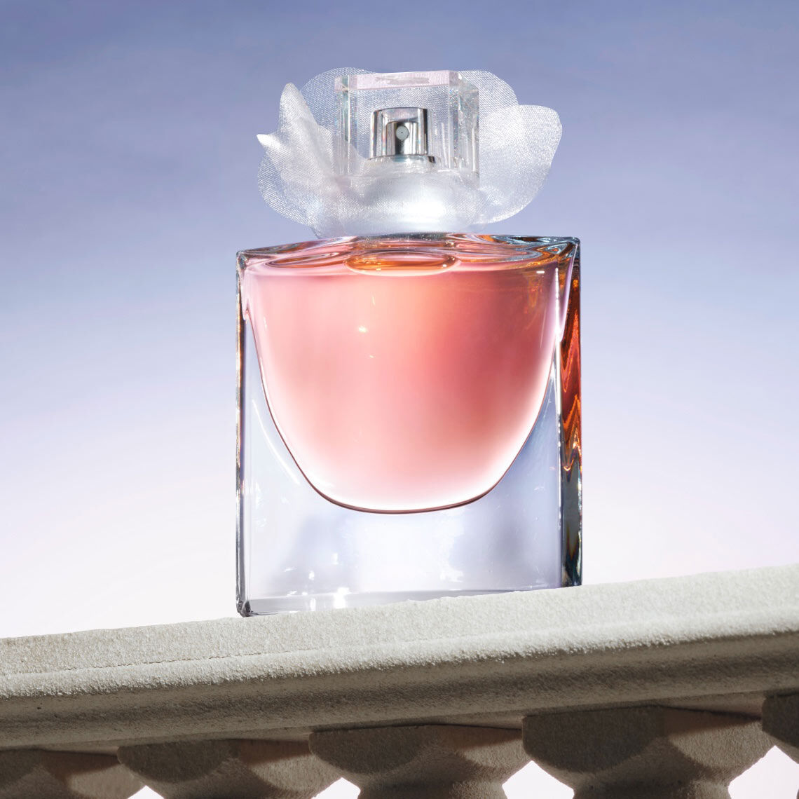 A clear glass perfume bottle with a soft pink liquid inside, topped with a delicate white flower-shaped cap, is displayed against a gradient background.