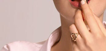 A woman with light brown hair applies lipstick while wearing a white button-up shirt and a gold ring. She is facing the camera and has a neutral expression.