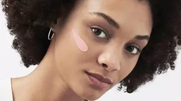 A young woman with curly hair is applying a pink skincare product to her cheek, looking directly at the camera with a soft expression.