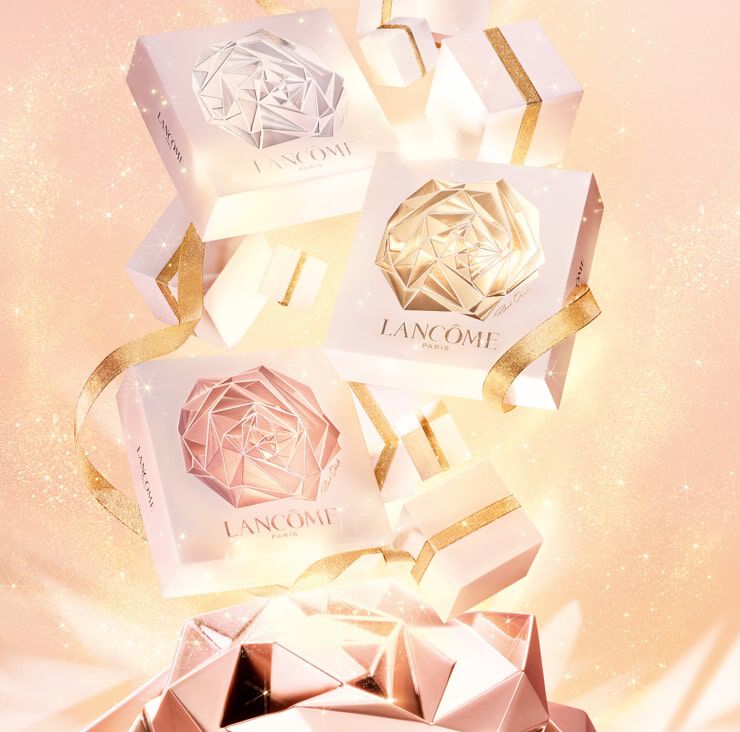 Three elegantly designed gift boxes in white and gold with a geometric rose motif, surrounded by smaller gift boxes and a sparkling background. The boxes are labeled "Lancome Paris."