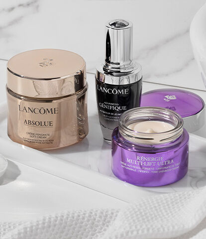 A clean, elegant bathroom countertop featuring three skincare products: a gold jar from Lancome, a black serum bottle, and a purple jar from another brand, arranged on a white tray with a cotton pad and a white towel in the background.