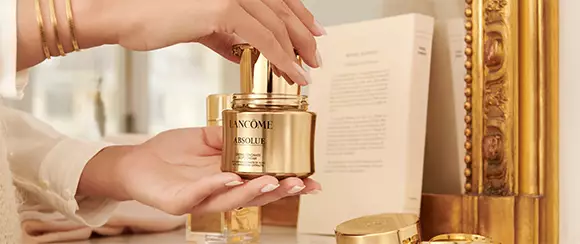 A close-up of a person's hands holding a luxurious gold jar of Lancome Absolue skincare product, with elegant decor in the background.
