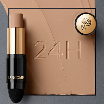 A Lancome foundation stick with a soft applicator next to a small compact, displayed on a background with swirls of foundation. The number "24H" is embossed in the surface.