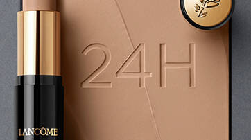 A Lancome foundation stick with a soft applicator next to a small compact, displayed on a background with swirls of foundation. The number "24H" is embossed in the surface.