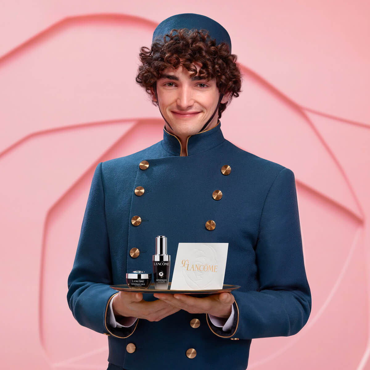 A smiling young man dressed in a blue uniform holds a tray featuring Lancome skincare products, set against a soft pink background.