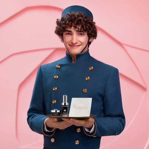 A smiling young man dressed in a blue uniform holds a tray featuring Lancome skincare products, set against a soft pink background.