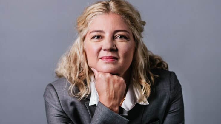 A woman with wavy blonde hair is resting her chin on her hand, dressed in a business suit, and looking confidently at the camera against a gray background.