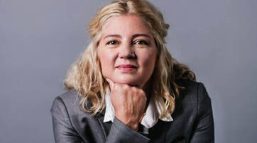 A woman with wavy blonde hair is resting her chin on her hand, dressed in a business suit, and looking confidently at the camera against a gray background.