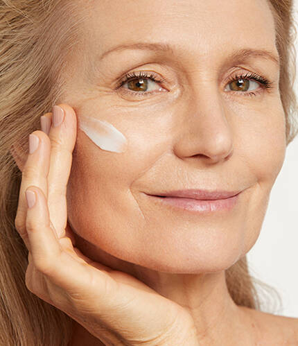 A woman with light hair gently applying cream to her cheek, smiling softly at the camera.