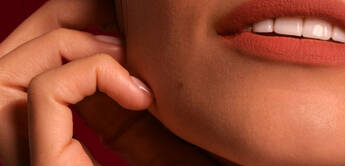 A close-up portrait of a woman with soft makeup, highlighting her features, and her hand gently touching her face against a dark red background.