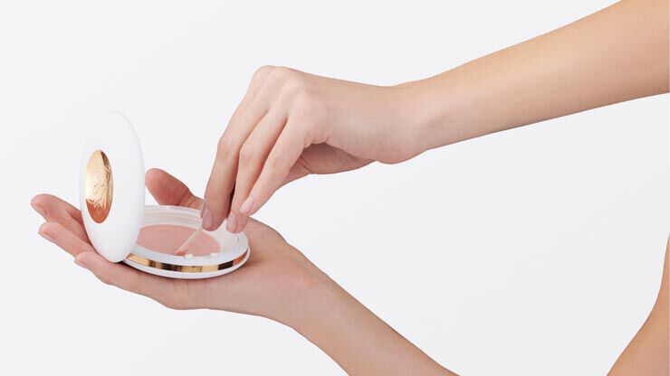 A hand holding a compact makeup case while another hand reaches inside to take some product. The compact has a sleek white design with a gold accent.