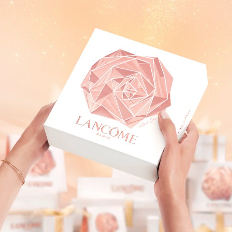 A person holding a beautifully designed white box with a geometric rose logo and the text "Lancome Paris" on it, set against a soft, sparkling background.