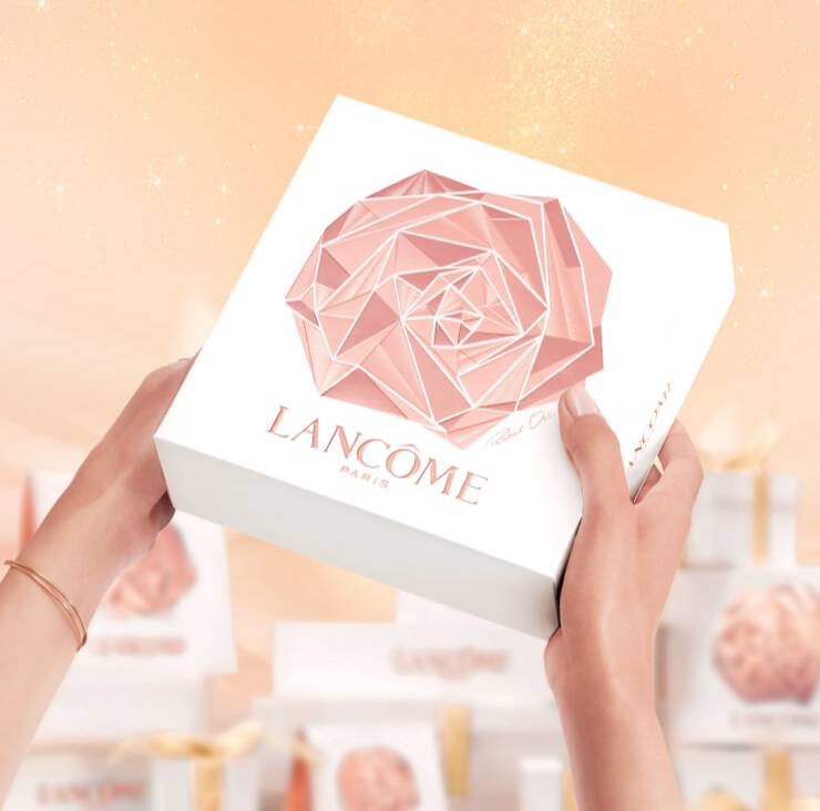 A person holding a beautifully designed white box with a geometric rose logo and the text "Lancome Paris" on it, set against a soft, sparkling background.